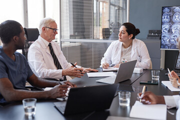 Middle Eastern female clinician writing key points of treatment plan on paper discussing anamnesis of patients disease with colleagues, while sitting at meeting table at medical office