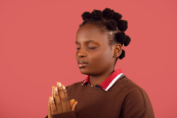 A serene portrait of a young woman with natural hair, in a praying pose with her hands together and eyes closed. Set against a calming coral backdrop, this image evokes peace and introspection.