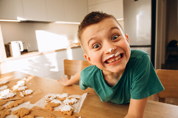 A school-age boy in the kitchen decorates Christmas cookies in sunny weather. Children in the kitchen, homemade pastries. Front view