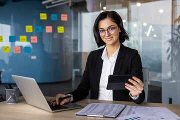 Confident businesswoman using laptop and smartphone for reports in office. Professional setting...