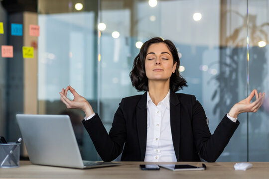 Woman in business attire meditating at office desk, eyes closed, embracing tranquility and mindfulness. Laptop and phone nearby emphasize balance between work and relaxation amidst workplace stress. - Powered by Adobe