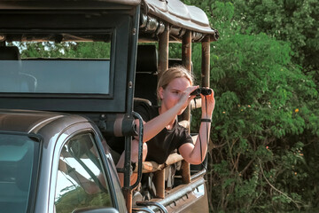 Woman in jeep on safari watching elephants with binoculars and capturing their beauty on camera. Concept of importance of wildlife conservation and eco-friendly travel in game reserves