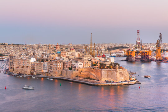 Sunset panoramic view of historic fortified city of Birgu (Vittariosa) with oil rig and other industrial facilities across the Grand Harbor from Valletta, Malta