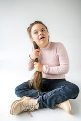 A young woman with Down syndrome sits with her legs crossed, wearing a pink sweater and blue jeans. She is looking up and smiling