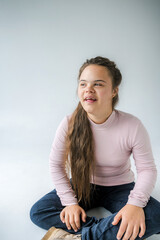 A young woman with Down syndrome sits with her legs crossed on a white background. She is wearing a pink long-sleeved shirt and blue jeans. She is looking to the right and smiling