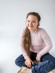A young woman with Down syndrome smiles confidently at the camera while sitting on the floor. She is wearing a pink turtleneck sweater and blue jeans