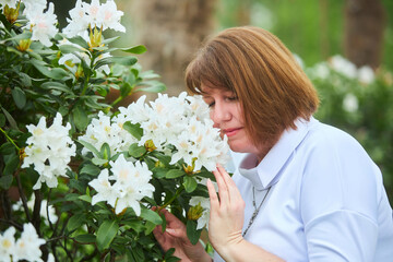 A middle-aged adult woman in a white dress stands near a bush blooming with white flowers.