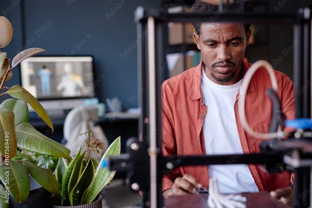 Leinwandbilder African American man working with 3D printer in modern office space focusing on precise details with computer in background and plants decorating workspace