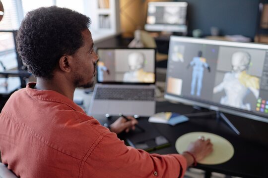 Man working on detailed 3D animation project at modern office desk, engrossed in development on dual monitors with laptop and other tools surrounding