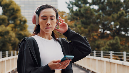 Japanese woman, headphones and phone in city of listening to music, fitness podcast and social media. Female athlete, mobile and streaming radio of exercise routine, training website and Japan bridge