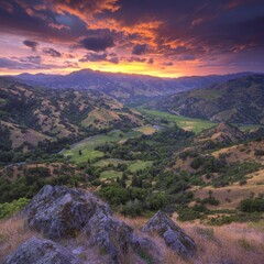 Vibrant sunset over a valley landscape with rolling hills, green meadows, and rocky foreground.