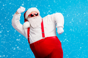 Photo of old man grey beard dance having fun raise hand fists wear santa claus x-mas costume cap headwear sunglass suspenders white shirt gloves isolated blue color background