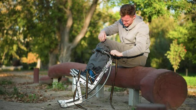 Young man sitting on reddish-brown log in park wearing light gray sweatshirt and khaki pants, focused on attaching straps to spring stilts beside him, surrounded by green trees
