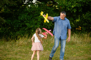 Fototapeta premium Happy girl and dad having fun outdoors, playing with toy airplane, running with it, in park on grass, smiling happily. Childhood and dream concept, father and child games. International Father's Day