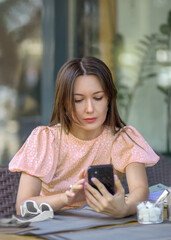 A young beautiful European woman with long hair holds a smartphone in her hands, checks social networks, uses a mobile application while sitting at an outdoor table in a cozy modern cafe.