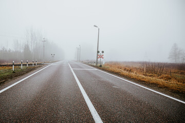 An empty rural road leading to a railroad crossing, surrounded by foggy fields and barren trees....