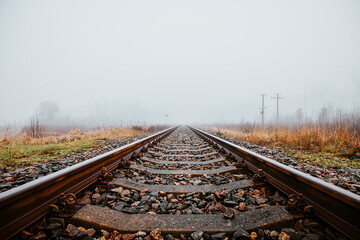 Fototapeta premium Straight railroad tracks surrounded by foggy fields and grass, leading into the horizon. The mist and muted colors evoke solitude and serenity.