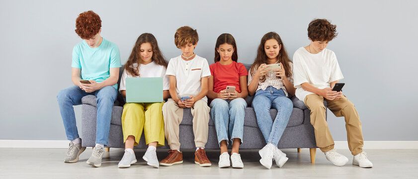 Group of young children school students sitting in a row on sofa together using mobile phones and laptop for social media isolated on gray wall background. Kids and technology concept.
