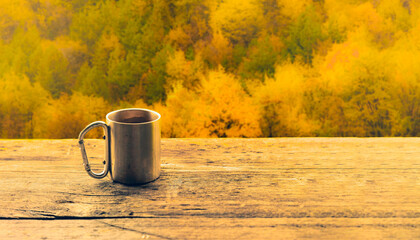 Stainless steel camping mug on a rustic wooden table with a vibrant autumn forest in the...