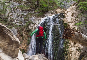 Obraz premium Hiker with trekking poles and a red backpack standing in front of a cascading waterfall surrounded by rocky cliffs and green foliage. A scene of outdoor adventure and natural beauty.