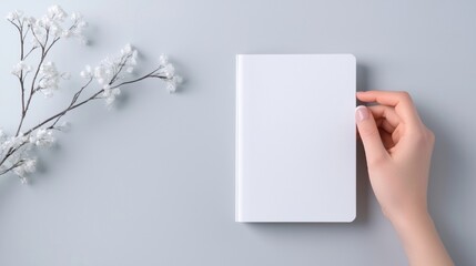 Obraz premium Hand holding a blank white notebook on a light blue background. on the left side of the image, there is a branch with small white flowers.