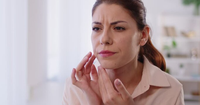 Close up of young woman standing in front of mirror, closely examining her face with concerned expression, dissatisfied with condition of her skin, noticing imperfections and signs of aging or stress.