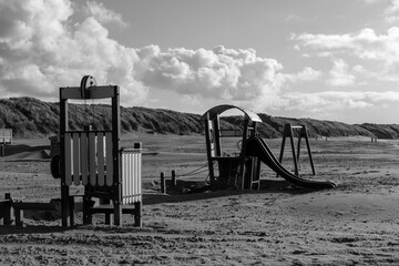 A black-and-white image of a deserted beach playground featuring swings and a slide, with sand dunes and dramatic clouds in the background under soft natural light.