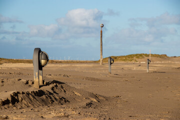 A sandy beach landscape with evenly spaced modern bollard lights partially buried in sand, distant...