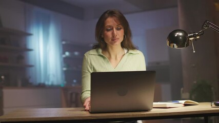 Attractive focused young woman sitting at desk and opening laptop for work in evening. Portrait of good-looking Caucasian girl working from home. Cozy apartment. Tracking moving around shot. Remote