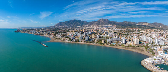Obraz premium Aerial view of Benalmadena, Spain, featuring white and pastel buildings, sandy beach, pier, turquoise sea, and lush hills under a clear blue sky.