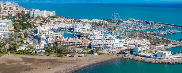 The image captures Benalmadena's marina with yachts, sandy beaches, and diverse architecture. A Ferris wheel stands near the turquoise Mediterranean Sea.