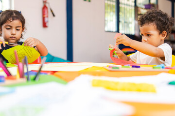 Latino boy and girl in preschool class cutting colorful papers at colorful tables during...