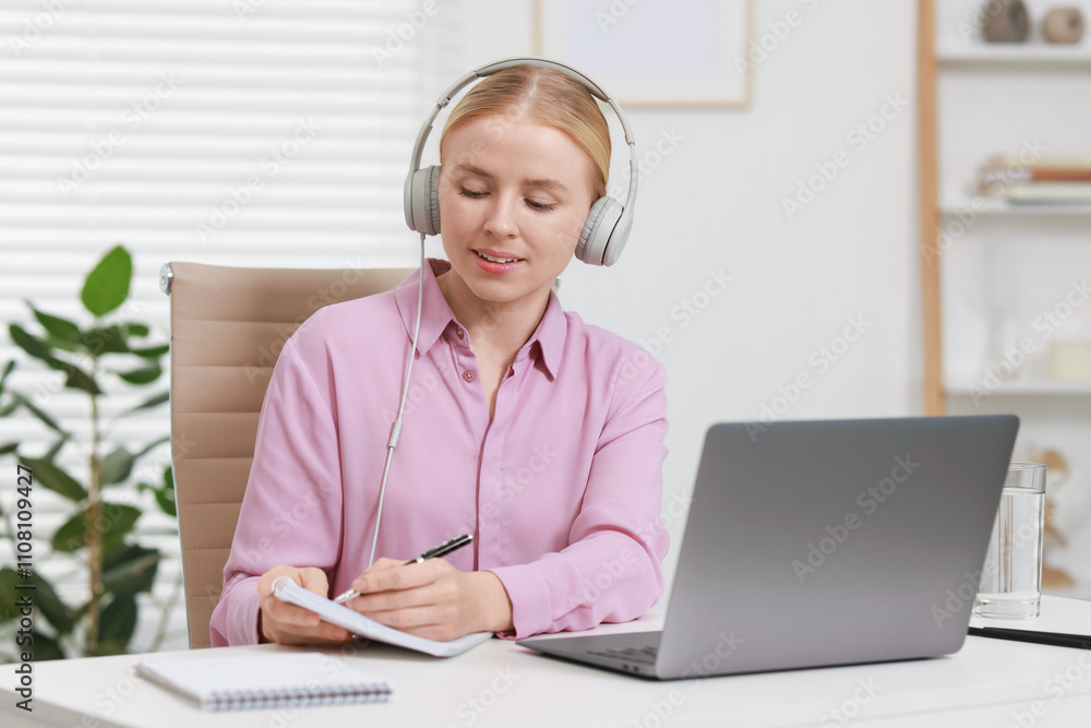Poster interpreter in headphones taking notes while having video chat via laptop at table indoors