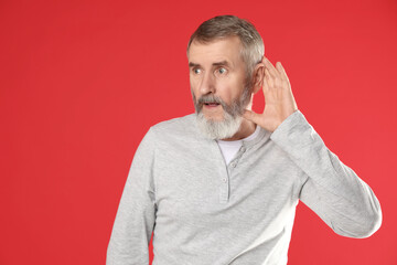 Senior man showing hand to ear gesture on red background
