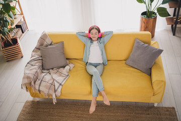Adorable girl in casual denim attire enjoying music with headphones while relaxing on a cozy yellow...