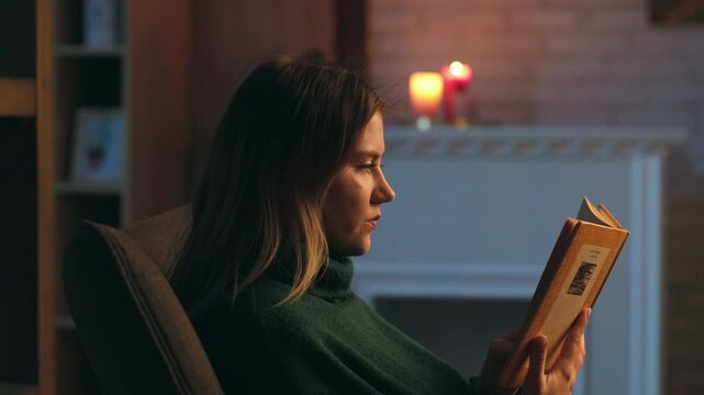 Close up young beautiful woman reading book sitting on sofa in the evening. Focused on novel. Relax at home