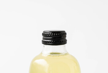 Small transparent glass bottle with black cap and white label, filled with colorful kombucha of yellow, standing on a white surface.