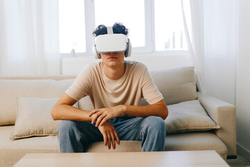 Virtual reality experience with a young man wearing a VR headset sitting on a beige couch in a bright room with large windows and soft curtains, conveying technology and leisure