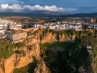 Obraz premium Ronda, Spain is seen from above at sunset, featuring the Puente Nuevo bridge spanning a dramatic gorge, with whitewashed buildings and a rugged landscape.