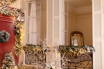 Snow covered balcony with wrought french iron railings with open shutters and doors. Old window with wrought-iron balcony decorated branches fir and glowing garland on Christmas	
