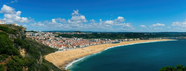 Obraz premium Breathtaking panoramic view of nazare beach, featuring golden sands, turquoise waters, and the charming cityscape nestled between the cliffs and the ocean, on a beautiful summer day in portugal