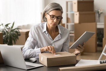 Busy grey haired e-commerce business owner woman checking barcode on paper package, using online...