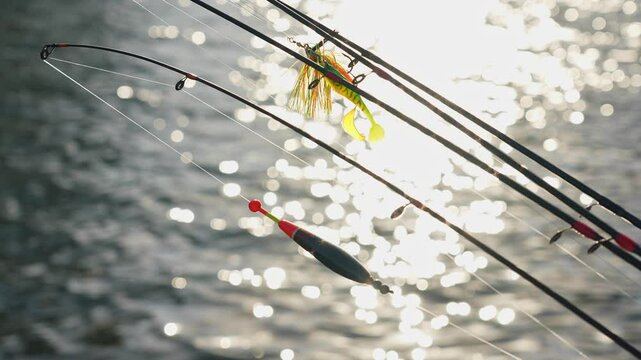 Fishing spinning and casting rods with fishing tackles on pier with blur lake background, selective focus. Fishing rods lines, bobber and plastic jig with worm lures. Slow motion movement on the wind.