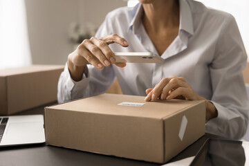 Mature e-commerce storage worker woman using mobile phone camera for reading barcode on cardboard box, parcel, paper package, preparing purchase for shipping from internet store