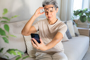 A middle-aged woman sits on the couch and lifts her glasses trying to read a message on the phone.