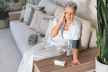 Mature beautiful middle-aged woman with gray hair sits on the couch and measures blood pressure