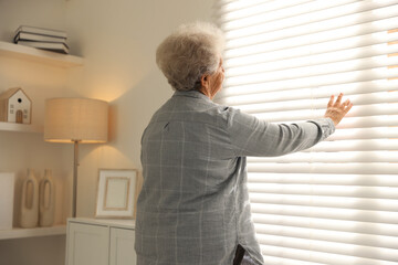 Fototapeta premium Loneliness concept. Sad senior woman looking through window blinds at home