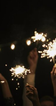 VERTICAL VIDEO: Burning sparklers in hands of group of friends on christmas. People waving new year lights in air on black background. Friends at party
