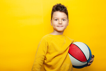 A boy wearing a bright yellow sweatshirt stands against a vivid yellow background, holding a colorful basketball with red, white, and blue sections. He has a calm, confident expression