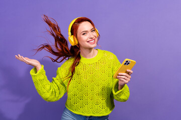 Cheerful young woman with red hair in green sweater listening to music on yellow headphones and...